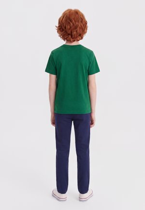 Enfant aux cheveux roux bouclés portant un t-shirt vert, un pantalon bleu marine et des baskets blanches, debout dos au spectateur sur un fond blanc.