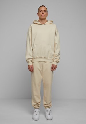 Beige hoodie met een voorzak en een capuchon met trekkoorden, gecombineerd met bijpassende sweatpants. De stof lijkt zacht met elastische boorden. Witte sneakers.
