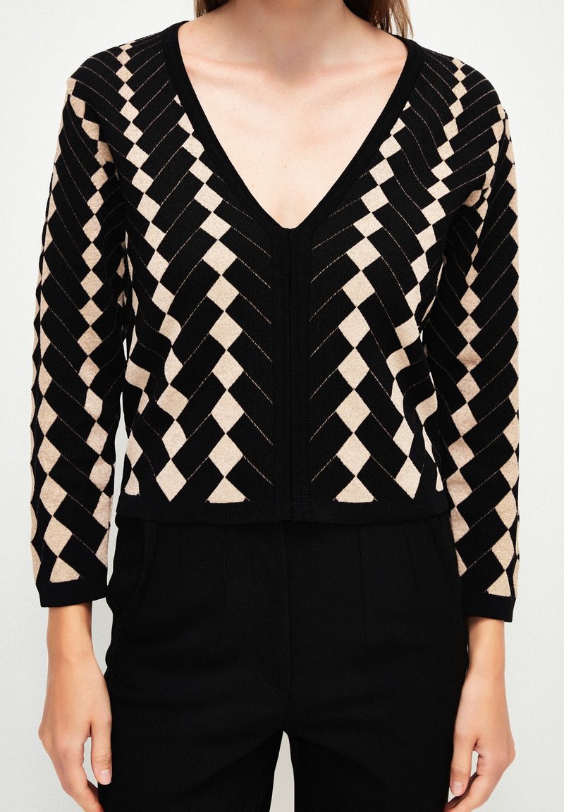 Haut à manches longues noir et beige avec un col en V, présentant un motif géométrique en chevron et en losange, associé à un pantalon noir.