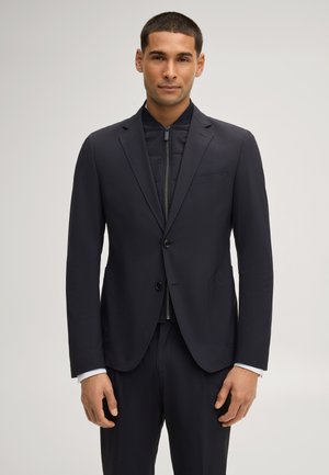 Man draagt een donker colbert over een zwart doorgestikt gewatteerd vest met rits, staand tegen een effen lichte achtergrond, recht vooruit kijkend.