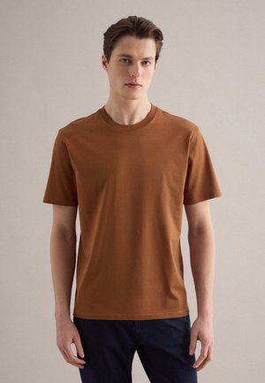 Giovane uomo in piedi che indossa una t-shirt marrone semplice a maniche corte e pantaloni scuri su uno sfondo neutro.