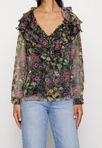 Blouse noire transparente avec un motif floral vibrant, dotée d'un col et de poignets volantés avec une bordure contrastante ; manches longues et coupe décontractée.