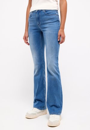 Person wearing light blue flare jeans, white sneakers, and a white top, standing against a plain white background.