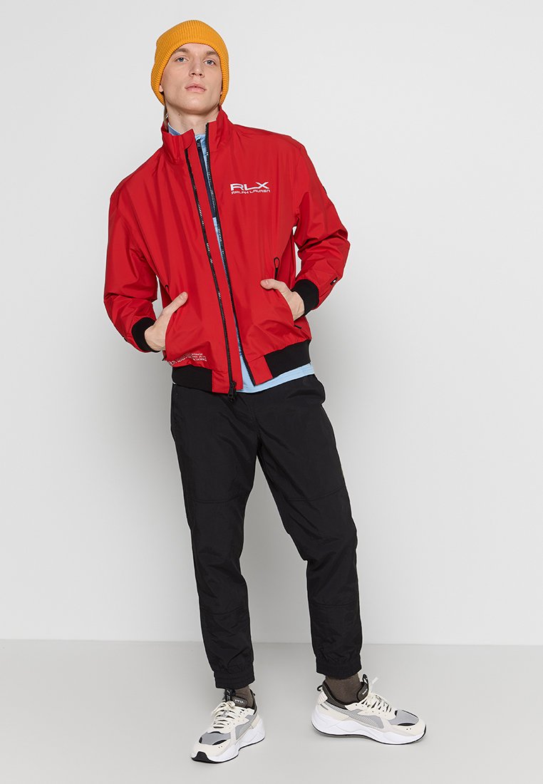 Chaqueta de nailon roja con cuello alto, cierre de cremallera y detalles en negro, combinada con pantalones negros y un gorro amarillo. Las zapatillas completan el conjunto.