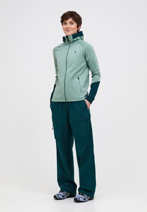 Persoon draagt een lichtgroene jas met ritssluiting en donker groene broek, met handen in de zakken, glimlacht en staat tegen een effen witte achtergrond.