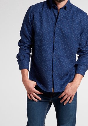Uomo che indossa una camicia a maniche lunghe blu scuro fantasia con bottoni, con le mani posate vicino ai fianchi, abbinata a jeans blu.