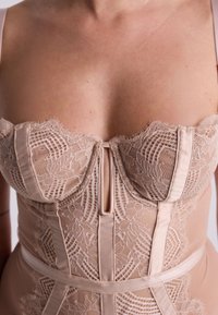 Corsetto beige con dettagli in pizzo, accenti in raso e una scollatura a chiave frontale. Presenta coppe strutturate e una vita aderente, esaltando la silhouette.