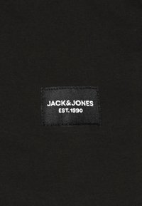 Fekete szövet, amelyen egy fekete címke van varrva, rajta a "JACK & JONES EST. 1990" felirattal fehér betűkkel. Sima textúra, látható minták nélkül.