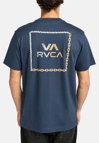Camiseta de algodón azul marina con un diseño gráfico de cadena y cuerda dorada, con el logo 'VA RVCA' centrado en la parte posterior. Manga corta, ajuste relajado.