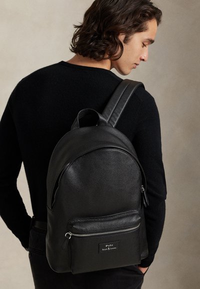 Uomo con maglione nero che porta uno zaino in pelle nera Polo Ralph Lauren con tasca frontale con zip, mostrato di spalle su sfondo neutro.