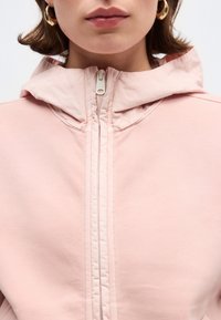 Κοντινό πλάνο ενός ατόμου που φοράει απαλό ροζ μπουφάν με κουκούλα και φερμουάρ, καθώς και χρυσούς κρίκους στα αυτιά, εμφανίζοντας το κάτω μέρος του προσώπου και τον λαιμό.