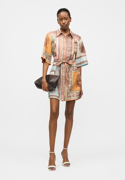 Γυναίκα που φοράει φόρεμα πουκάμισο με ζώνη και σχέδιο, με κοντά μανίκια, κρατώντας ένα σκούρο καφέ φάκελο και άσπρα πέδιλα με τακούνια.