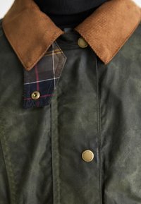 Πράσινο αδιάβροχο βαμβακερό μπουφάν με καφέ χνουδωτό γιακά, με καρό σχέδιο κάτω από τον γιακά και λεπτομέρειες από μπρούτζο στις κουμπωτές κλείσεις.