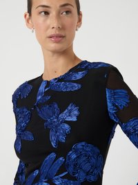 Kvinna som bär en svart genomskinlig långärmad topp med texturerad kungligt blå blom- och fågelbroderi, vänd något åt vänster, neutral bakgrund.