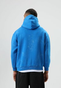 Sweat-shirt bleu à capuche, présentant une texture usée et des motifs légèrement plus clairs subtils. Le dos montre une coupe décontractée et un design de poignets typique.