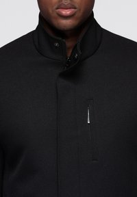 Zwarte jas gemaakt van gestructureerde stof met een hoge kraag, voorzien van een ritssluiting aan de voorkant en een klein ritssluitvak aan de linkerkant.