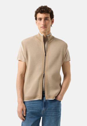 Jeune homme portant une veste beige sans manches zippée par-dessus une chemise à manches courtes, associée à un jean bleu, debout devant un fond uni.