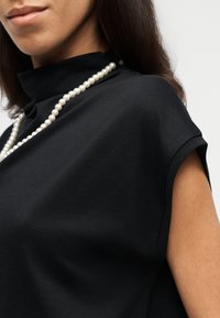 Prim-plan cu o persoană care poartă un top negru fără mâneci, cu guler înalt, și un colier simplu de perle, pe un fundal deschis și simplu.