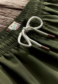 Rohelised ujumispüksid elastse vöökohaga, valge pitsikujundusega, mille otstes pruunid narmad ning silt, millel on palmi puud kujutavad graafikud.