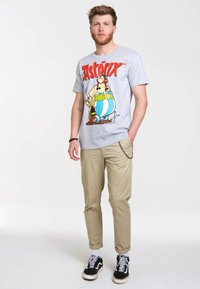 Graues T-Shirt mit Asterix-Charaktergrafik in Rot und Gelb. Kombiniert mit beigen Hosen und schwarzen Sneakers mit weißen Socken.