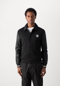 Sort varsity-jakke med en ensfarvet stofkrop og læderærmer, der har et logo på brystet og stribede ribbede manchetter.