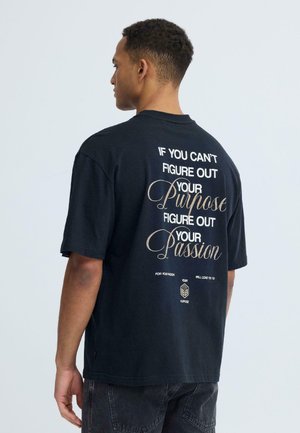 Mand set bagfra iført en sort t-shirt med hvid og beige tekst: "Hvis du ikke kan finde ud af dit formål, så find ud af din passion."