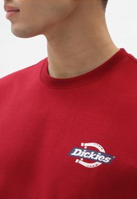 Sudadera de algodón roja con cuello redondo y un parche de logo etiquetado "Dickies" en azul, con acentos en blanco y un gráfico de herradura.