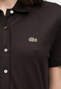 Polo noir à manches courtes avec des boutons blancs et un petit logo de crocodile vert sur le côté gauche de la poitrine.