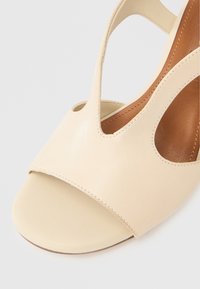 Μπεζ δερμάτινο σανδάλι με ανοιχτό δάχτυλο και Τ-λαιμόκοψη με ορατές ραφές, διαθέτει καφετί μαξιλαρωμένη εσωτερική σόλα πάνω σε λευκό φόντο.