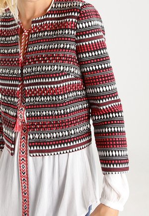 Veste courte avec des motifs géométriques rouges, noirs et blancs intriqués. Le tissu présente une broderie texturée et un col rond.