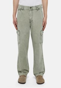 Muž v bledě zelených cargo kalhotách a béžových nazouvacích botách, stojící s uvolněnýma rukama po stranách na jednobarevném pozadí.