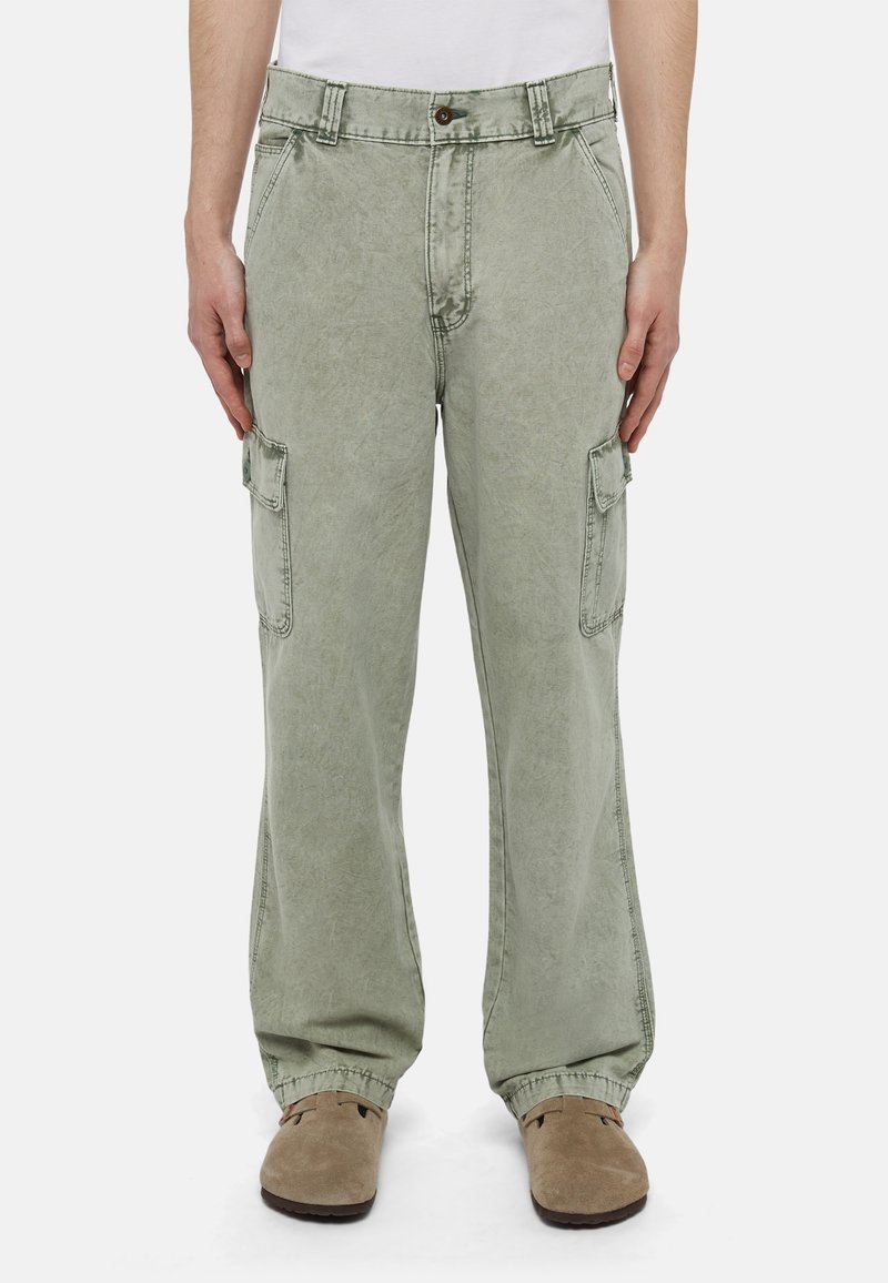 Muž v bledě zelených cargo kalhotách a béžových nazouvacích botách, stojící s uvolněnýma rukama po stranách na jednobarevném pozadí.