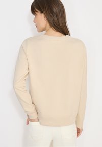 Femme aux cheveux bruns raides portant un pull beige à manches longues et un pantalon blanc, vue de dos.