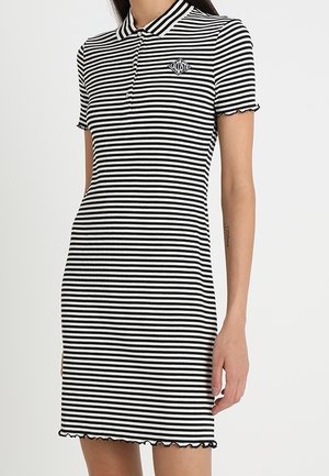 Kobieta ubrana w czarno-białą sukienkę polo w poziome paski z krótkimi rękawami i małym logo Lacoste na piersi.