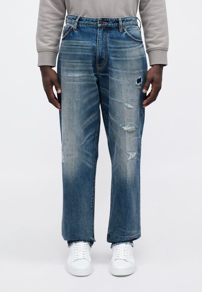 Jeans de mezclilla azul claro con un corte holgado, detalles desgastados y decoloración en todo el pantalón. Combinados con zapatillas blancas.