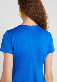 Blå kortärmad klänning med en mjuk textur, rund halsringning och dragkedja i ryggen. Subtil sömdetalj går vertikalt längs ryggen.