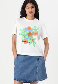 Bela bombažna majica s pisanimi grafičnimi vzorci, ki vključujejo zelene palmove liste, oranžne kroge in besedilo "Monaco". V kombinaciji z modrim denim krilom.