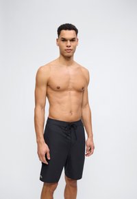 Άνδρας χωρίς μπλούζα που φοράει μαύρο μαγιό με κορδόνι, στέκεται μπροστά σε απλό φωτεινό φόντο, κοιτάζοντας προς τα εμπρός.