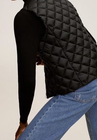 Černá prošívaná vatovaná vesta s vysokým límcem, kombinovaná s černým přiléhavým tričkem s dlouhým rukávem a modrými džíny, ukazující texturované vzory a švy.