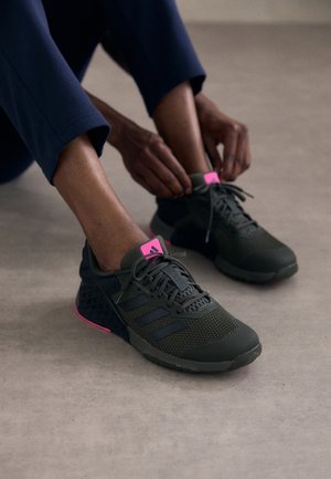 Person in marineblauer Hose bindet die Schnürsenkel schwarzer Sneakers mit pinken Akzenten, während sie auf einem grauen Boden sitzt.