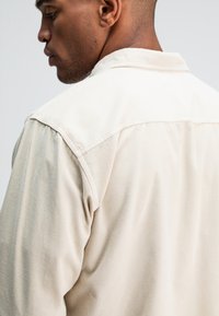 Beige corduroy overhemd met een gestructureerde kraag en gedetailleerde stiksels, die textuur en stofgranulatie vanuit een zijaanzicht toont.