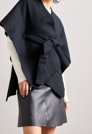 Vrouw die een zwarte gebreide sjaal draagt, geknoopt aan de voorkant, over een witte geribbelde top met lange mouwen en een zwarte leren rok.