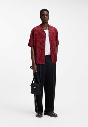 Hombre con camisa roja de manga corta con flores, camiseta blanca interior, pantalones anchos negros, zapatos negros, sosteniendo una bolsa negra Hugo sobre fondo blanco.