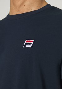 Granatowa bawełniana koszulka z okrągłym dekoltem i małym, haftowanym logo w kolorze czerwonym, białym i niebieskim po lewej stronie klatki piersiowej.