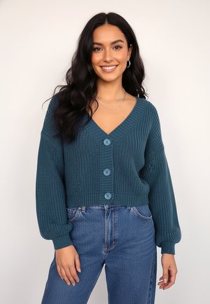 Morski, dziergany sweter na guziki z dekoltem w serek, czterema niebieskimi guzikami i bufiastymi rękawami, zestawiony z wysokim stanem dżinsów. Widoczny detal tekstury tkaniny.