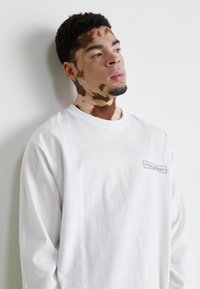 Weißes Langarm-T-Shirt aus Baumwolle mit Rundhalsausschnitt und kleinem, gesticktem Logo auf der linken Brust. Glatte Textur.