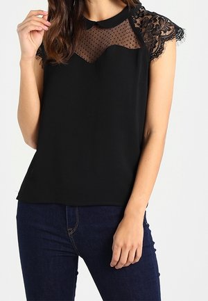 Mujer que lleva una camiseta negra sin mangas con mangas cortas de encaje y canesú transparente con topos, combinada con vaqueros azul oscuro.