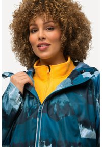 Chaqueta verde azulado con patrones abstractos en azul oscuro, con cierre frontal y capucha ajustable, superpuesta a una prenda interior de forro polar amarilla.