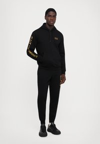Zwarte hoodie en broek set, met gele branding op de mouwen en dij, gemaakt van zachte stof met een relaxte pasvorm en kangoeroezak.