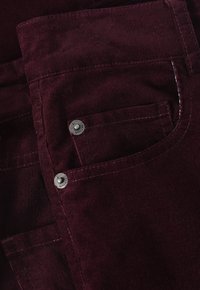 Pantalon en velours côtelé bordeaux avec une texture lisse, présentant des boutons en métal argenté sur la poche avant et des détails de couture contrastants.